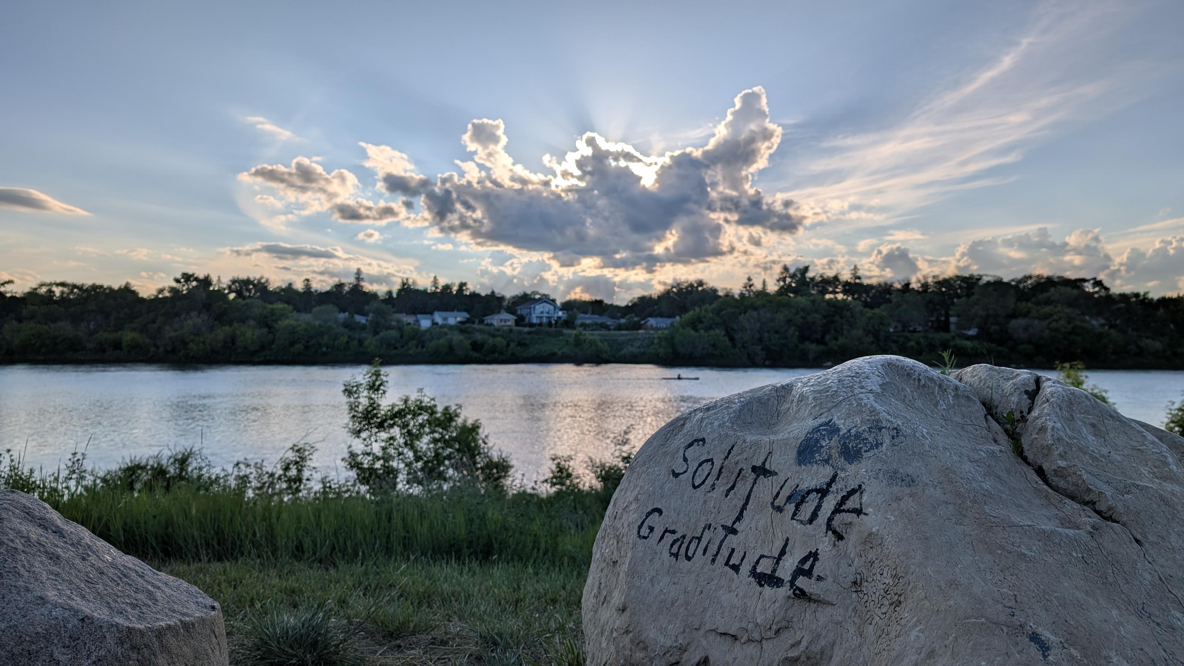 A nice skyline with a river has a cloud with "God-rays" behind it above. A Rock in the foreground says "Solitude, Gratitude" on it.