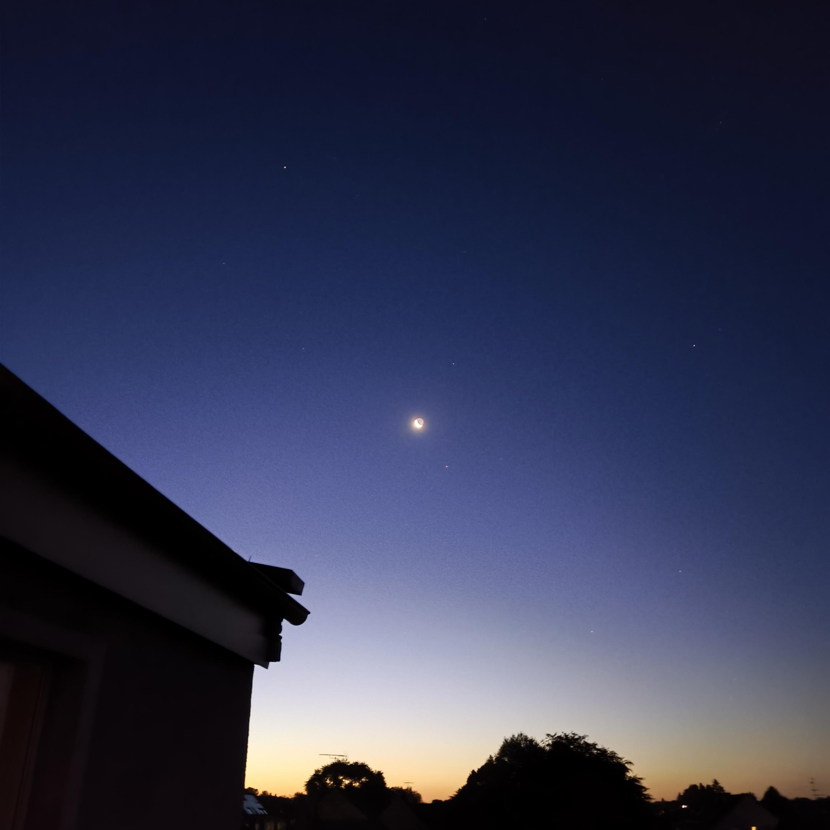 Picture of the cresent moon (in the centre) and the night sky before sunrise. It shows a gradient from light yellow, light purple to dark blue. At the bottom of the picture, he black shillouettes of some trees and houses are visible.